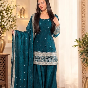 Puede incluir: Un conjunto de salwar kameez color verde azulado que incluye una túnica de manga larga con bordados plateados en el dobladillo y los puños, pantalones anchos a juego y un dupatta. La tela tiene un brillo sutil y pequeños puntos plateados.