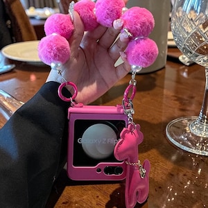 Puede incluir: Una funda de teléfono rosa para un teléfono Galaxy Z Flip, con un charm de pompón rosa a juego y un llavero con forma de perro rosa. La funda del teléfono tiene la inscripción "Galaxy Z Flip".