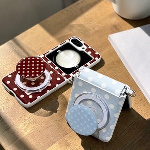 May include: Two phone cases with polka dot designs. One case is burgundy with white dots, and the other is light blue with white dots. Both cases have a circular pop-up grip with a matching polka dot pattern. The phone cases are on a wooden surface.