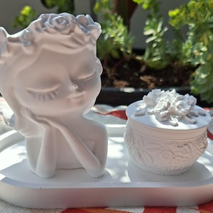 Könnte beinhalten: Ein weißes dekoratives Keramik-Set mit einer Büste einer Frau als Pflanzgefäß mit Blumenkranz und einer kleinen runden Dose mit Deckel und floralen Details. Das Set steht auf einem weißen ovalen Tablett, geeignet für kleine Pflanzen oder Schmuck.