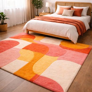 Op de afbeelding: Een kleurrijk vloerkleed met een geometrisch ontwerp in tinten oranje, roze en crème. Het rechthoekige kleed ligt op een houten vloer in een slaapkamer. De kamer heeft ook een bed met kussens en een geweven hoofdbord.