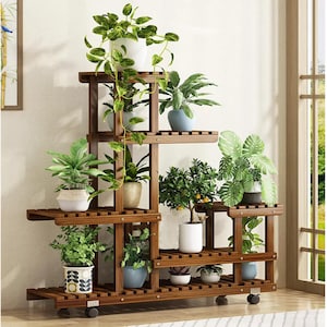 May include: A multi-tiered, dark brown wooden plant stand with multiple shelves. The stand is filled with various potted plants, including leafy green plants and small trees. The stand has wheels for easy movement and is placed near a window.