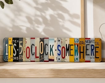 May include: A rectangular wall sign composed of various vintage-style license plates arranged horizontally. The plates feature colorful letters and numbers that spell out the phrase 1TS 5 OCLOCK SOMEWHERE in a mix of blue, red, black, and yellow.