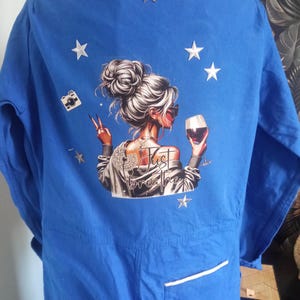 Puede incluir: Mono azul con un gráfico en la espalda que presenta a una mujer con gafas de sol sosteniendo una copa de vino y haciendo el signo de la paz. El texto "Just Breathe" está debajo de la mujer. Estrellas plateadas y un gráfico de cámara también están presentes.