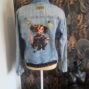 Peut inclure: Veste en jean bleu clair avec une bordure en dentelle noire. Le dos présente un graphisme d'une femme portant des lunettes de soleil, tenant un verre de vin, et le texte "SELF-MADE". Décorée d'étoiles et d'éclaboussures.
