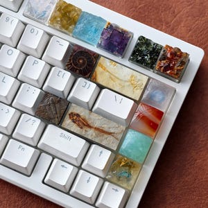 Könnte beinhalten: Eine weiße Tastatur mit individuellen Tastenkappen, die verschiedene Edelsteine, Fossilien und Mineralien zeigen. Die Tastenkappen weisen eine Reihe von Farben auf, darunter Blau, Gelb, Lila, Grün und Orange. Die Tastatur liegt auf einer braunen Lederoberfläche.