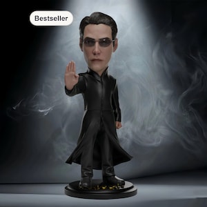 Puede incluir: Figura de bobblehead de un hombre con abrigo negro largo y gafas de sol, con la mano derecha levantada. La figura está sobre una base negra con detalles dorados. La palabra "Bestseller" está en un óvalo blanco.