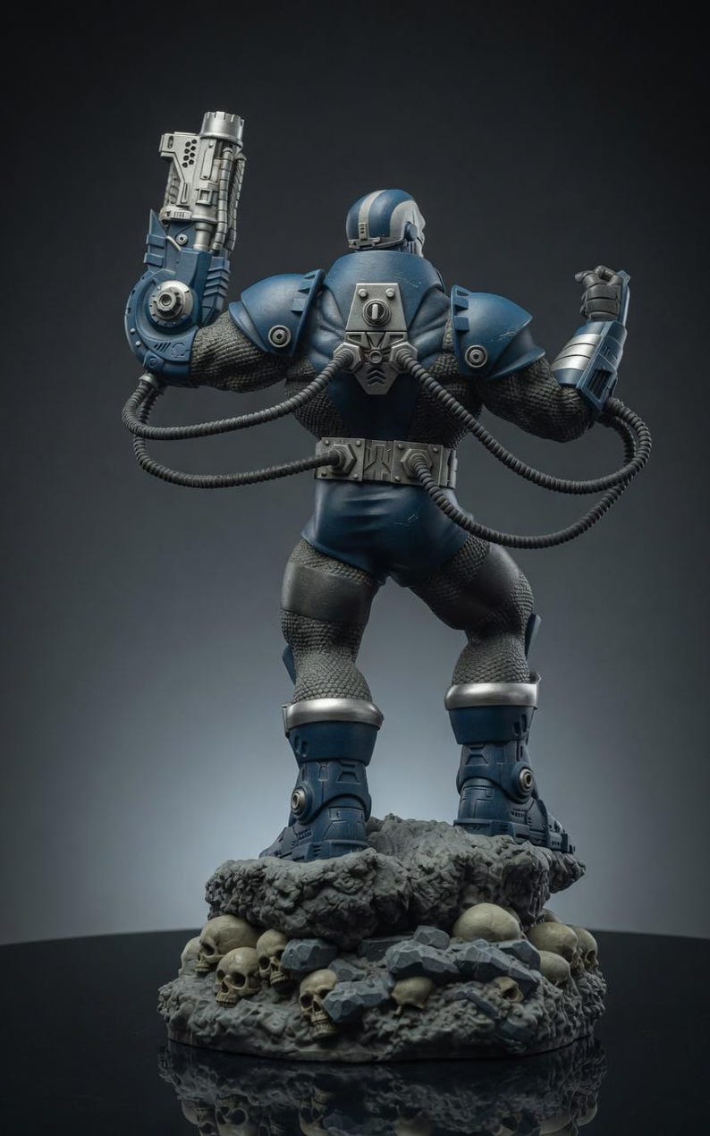 May include: A detailed statue of a blue and silver armored figure, standing on a rocky base with skulls. The figure has a mechanical arm and various tubes connected to its body. The statue is a collectible item.