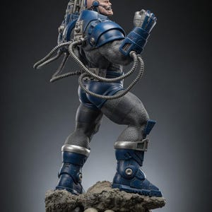 May include: A detailed statue of a muscular, blue-armored figure with a bald head and a grimacing expression. The figure stands on a rocky base with skulls, and has a large weapon on its back. The statue is a collectible item.