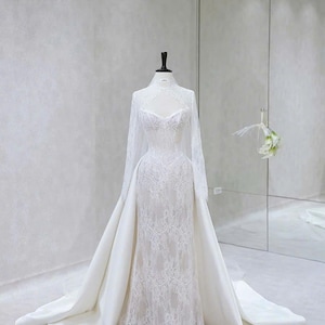 Könnte beinhalten: Ein bodenlanges weißes Brautkleid mit einem hochgeschlossenen Spitzenoberteil mit langen Ärmeln, einem herzförmigen Ausschnitt und einem abnehmbaren Satin-Überrock, der eine dramatische Schleppe bildet. Die aufwendige Spitze bedeckt das gesamte Kleid.