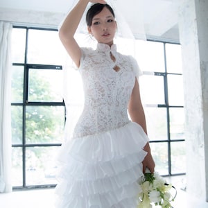 May include: A white lace wedding dress with a high neckline and short sleeves. The dress features a fitted bodice and a tiered, ruffled skirt. The bride is holding a bouquet of white flowers and wearing a veil and gloves.