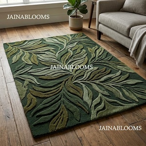 May include: A rectangular green rug featuring a leaf design in various shades of green. The rug is positioned on a wooden floor, with a sofa and a potted plant in the background. The text "JAINABLOOMS" is visible on the rug.
