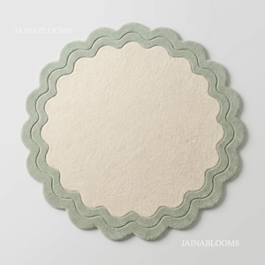 May include: A circular rug featuring a cream-colored center and a scalloped sage green border. The design includes a wavy, layered edge detail that frames the inner circle. The text JAINABLOOMS is visible in the top left and bottom right corners.