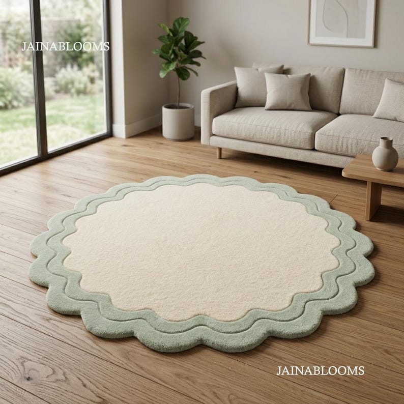 May include: A round area rug featuring a solid cream-colored center and a wide, scalloped sage green border. The rug has a soft, textured pile and a distinct wavy edge design that creates a layered appearance on the wooden floor surface.