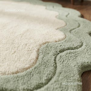 May include: A plush area rug featuring a cream-colored center and a wide, sage green scalloped border. The rug has a thick, tufted texture throughout. The text JAINABLOOMS is visible in the upper left and lower right corners of the image.