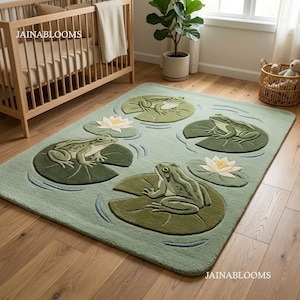 Op de afbeelding: Een groen vloerkleed met een vijverscène met kikkers op waterlelies en witte waterlelies. Het kleed heeft een lichtgroene achtergrond met donkergroene waterlelies en kikkers. De tekst "JAINABLOOMS" is zichtbaar.