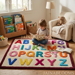 May include: A rectangular area rug featuring a cream-colored background with a dark red border and large, multi-colored, three-dimensional tufted letters of the alphabet arranged in rows. The rug is made of a soft, textured material.