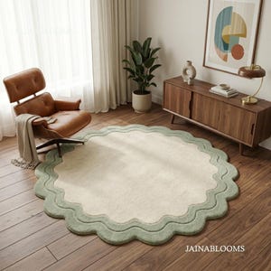 May include: A round area rug featuring a cream-colored center and a wide, scalloped sage green border. The rug is placed on a wooden floor in a room with a brown leather lounge chair, a wooden sideboard, and a potted plant. The text JAINABLOOMS is visible.