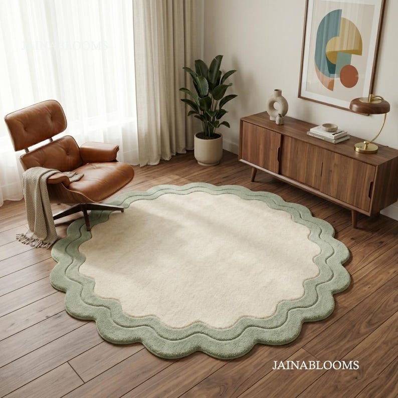 May include: A round area rug featuring a cream-colored center and a wide, scalloped sage green border. The rug is placed on a wooden floor in a room with a brown leather lounge chair, a wooden sideboard, and a potted plant. The text JAINABLOOMS is visible.