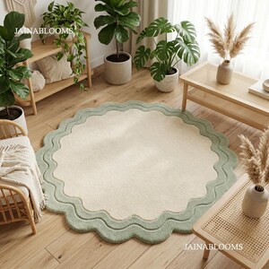 May include: A round area rug featuring a solid cream-colored center and a wide, scalloped sage green border. The rug has a soft, textured surface and is positioned on a light wood floor. The text JAINABLOOMS is visible in the bottom right corner.