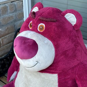 May include: A large, plush, magenta-colored bear with a white belly and snout. The bear has brown eyebrows, brown eyes with orange irises, and a pink nose. The bear's ears have white inner linings. The bear is a character from the Toy Story franchise.