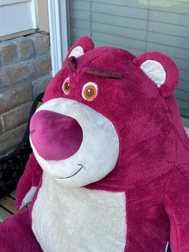May include: A large, plush, magenta-colored bear with a white belly and snout. The bear has brown eyebrows, brown eyes with orange irises, and a pink nose. The bear's ears have white inner linings. The bear is a character from the Toy Story franchise.