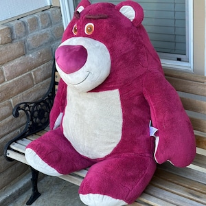May include: A large, plush, magenta-colored bear with a white belly and accents. The bear has a smiling expression with a pink nose and brown eyes. It is sitting on a wooden bench.
