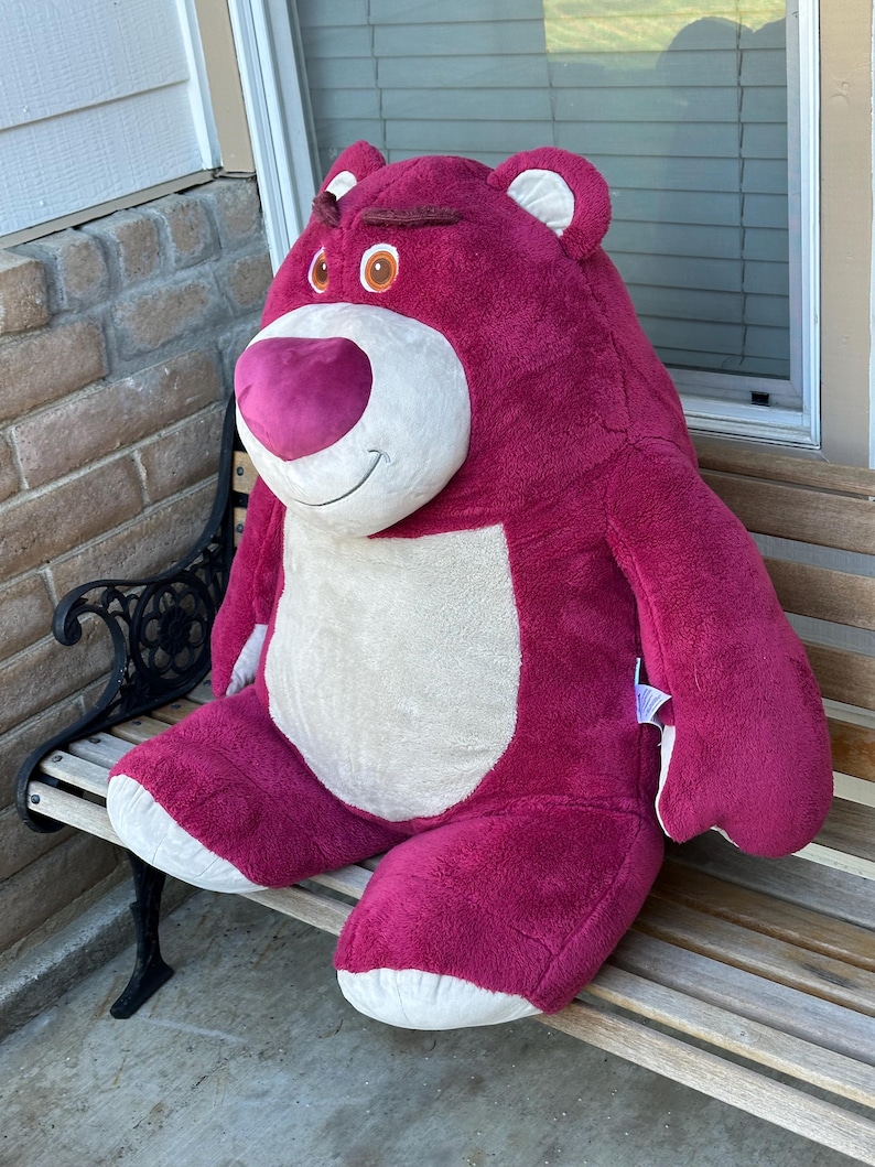 May include: A large, plush, magenta-colored bear with a white belly and accents. The bear has a smiling expression with a pink nose and brown eyes. It is sitting on a wooden bench.