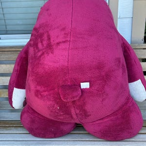 May include: A large, plush, magenta-colored stuffed animal with white accents on the paws. The toy is sitting on a wooden bench. The toy has a small tail and a white tag.