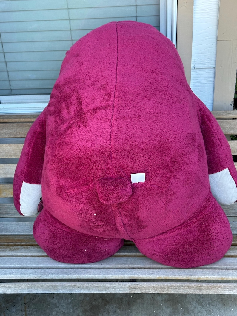 May include: A large, plush, magenta-colored stuffed animal with white accents on the paws. The toy is sitting on a wooden bench. The toy has a small tail and a white tag.