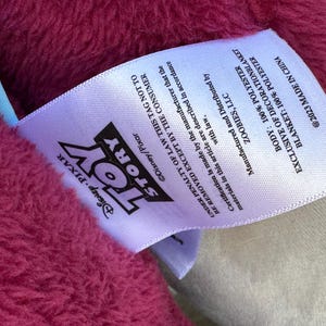 May include: A close-up of a pink blanket with a white tag. The tag features the "Toy Story" logo and text indicating it's made of 100% polyester. The blanket is exclusive of decoration and made in China.