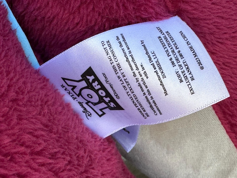 May include: A close-up of a pink blanket with a white tag. The tag features the "Toy Story" logo and text indicating it's made of 100% polyester. The blanket is exclusive of decoration and made in China.