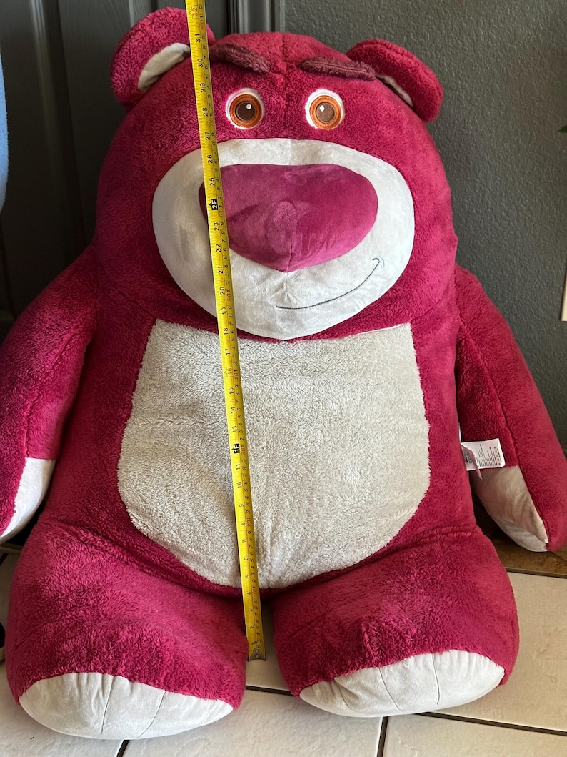 May include: A large, plush, pink teddy bear with a white belly and a heart-shaped nose. The bear is approximately 30 inches tall. It has a grumpy expression and is a character from a popular animated film.