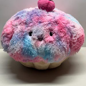 May include: A plush cupcake-shaped pillow with a smiling face. The pillow features a fluffy, tie-dye design in pink, blue, and purple. A pink cherry with a brown stem sits on top. The base of the cupcake is a light tan color.