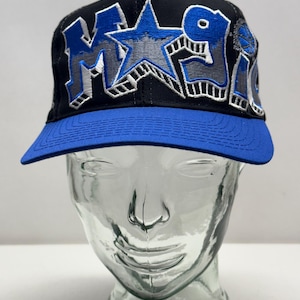 May include: Black and blue baseball cap with the word "MAGIC" embroidered in blue and grey. The cap has a blue brim and a star graphic. The hat is displayed on a glass head.