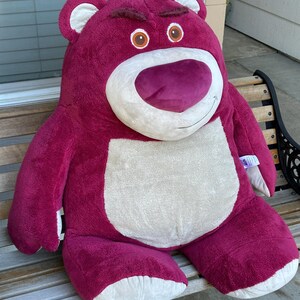 May include: A large, plush, magenta-colored teddy bear with a white belly and snout, and brown eyes. The bear has a stern expression and is sitting on a wooden bench. The bear's ears and feet are white.