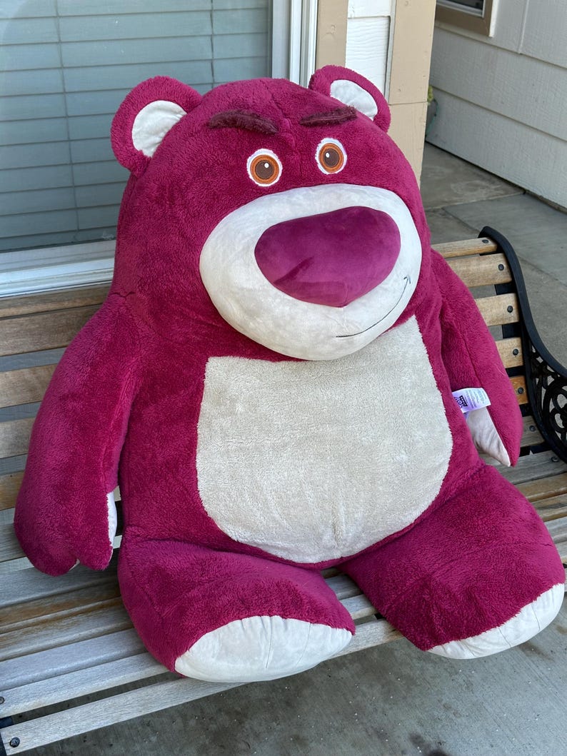 May include: A large, plush, magenta-colored teddy bear with a white belly and snout, and brown eyes. The bear has a stern expression and is sitting on a wooden bench. The bear's ears and feet are white.