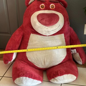May include: A large, plush red bear with a white belly and face, and brown eyes. The bear has a stern expression. A yellow measuring tape is stretched across the bear's body, indicating its size.