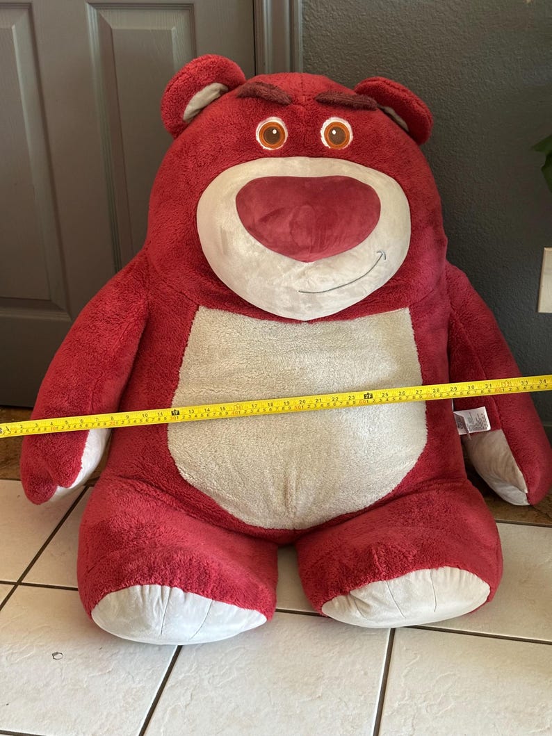 May include: A large, plush red bear with a white belly and face, and brown eyes. The bear has a stern expression. A yellow measuring tape is stretched across the bear's body, indicating its size.
