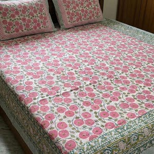 Può includere: Un copriletto in cotone bianco e due federe abbinate con un motivo ripetuto di fiori circolari rosa con steli e foglie verdi, rifinito con un bordo decorativo a righe verdi e bianche lungo i bordi del tessuto.