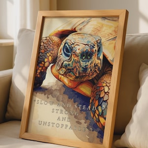 May include: A framed print featuring a colorful, stylized illustration of a tortoise head and neck. The artwork includes the text, "SLOW AND STEADY STRONG AND UNSTOPPABLE..." in a serif font. The frame is made of light-colored wood with a grain texture.