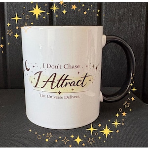 Könnte beinhalten: Weißer Keramikbecher mit schwarzem Henkel. Der Becher zeigt den Text "I Don't Chase, I Attract" in einer Schrift, darunter "The Universe Delivers". Das Design beinhaltet himmlische Akzente.