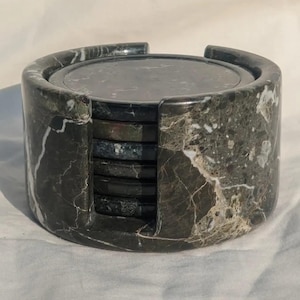 May include: A set of six dark green marble coasters with white veining, stored in a matching cylindrical holder. The coasters are round and have a polished finish, perfect for protecting surfaces from drink rings.