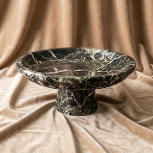 Puede incluir: Un cuenco circular con pedestal fabricado en mármol verde oscuro con vetas blancas prominentes. El cuenco presenta una base ancha y poco profunda sostenida por una base cilíndrica gruesa, mostrando la textura natural de la piedra pulida.