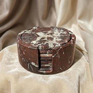 May include: A set of six marble coasters in a holder. The coasters and holder are a deep red with white and grey veining. The coasters are round and fit snugly in the matching holder, which has a cylindrical shape.