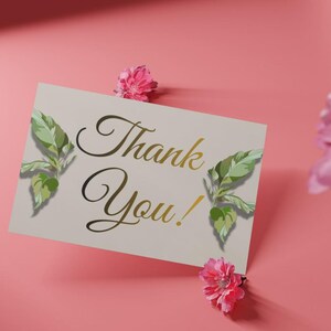 Op de afbeelding: Een crèmekleurige kaart met gouden belettering "Thank You!", versierd met groene bladeren en roze bloemen, tegen een roze achtergrond.