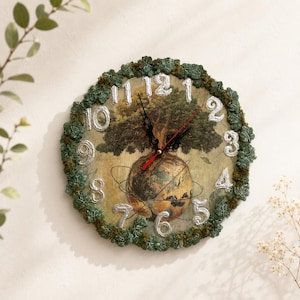 May include: A round wall clock featuring a central illustration of a tree growing from a globe. The clock face has silver-colored numbers and is framed by a textured border of green leaves and moss. It includes black hour and minute hands.