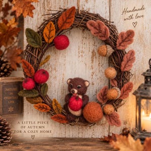 Pode incluir: Uma guirlanda circular feita de galhos castanhos decorada com elementos de lã feltrada, incluindo folhas laranja e castanhas, maçãs vermelhas e bolotas bege. Um pequeno urso de feltro castanho está sentado no centro segurando uma maçã.