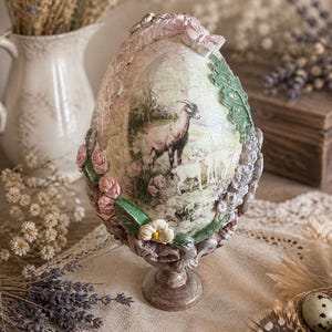 Può includere: Un oggetto decorativo a forma di uovo ornato con una scena pastorale raffigurante una capra e un agnello. L'uovo è decorato con abbellimenti floreali e fogliari nei toni del rosa, verde e crema, e poggia su un piedistallo decorativo.
