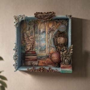 May include: A decorative shadow box with a fantasy theme. The box features a detailed interior scene with winged lion statues, books, and a globe. The frame is ornate with a light blue and brown color scheme, and the text "BE HAPPY" is visible.